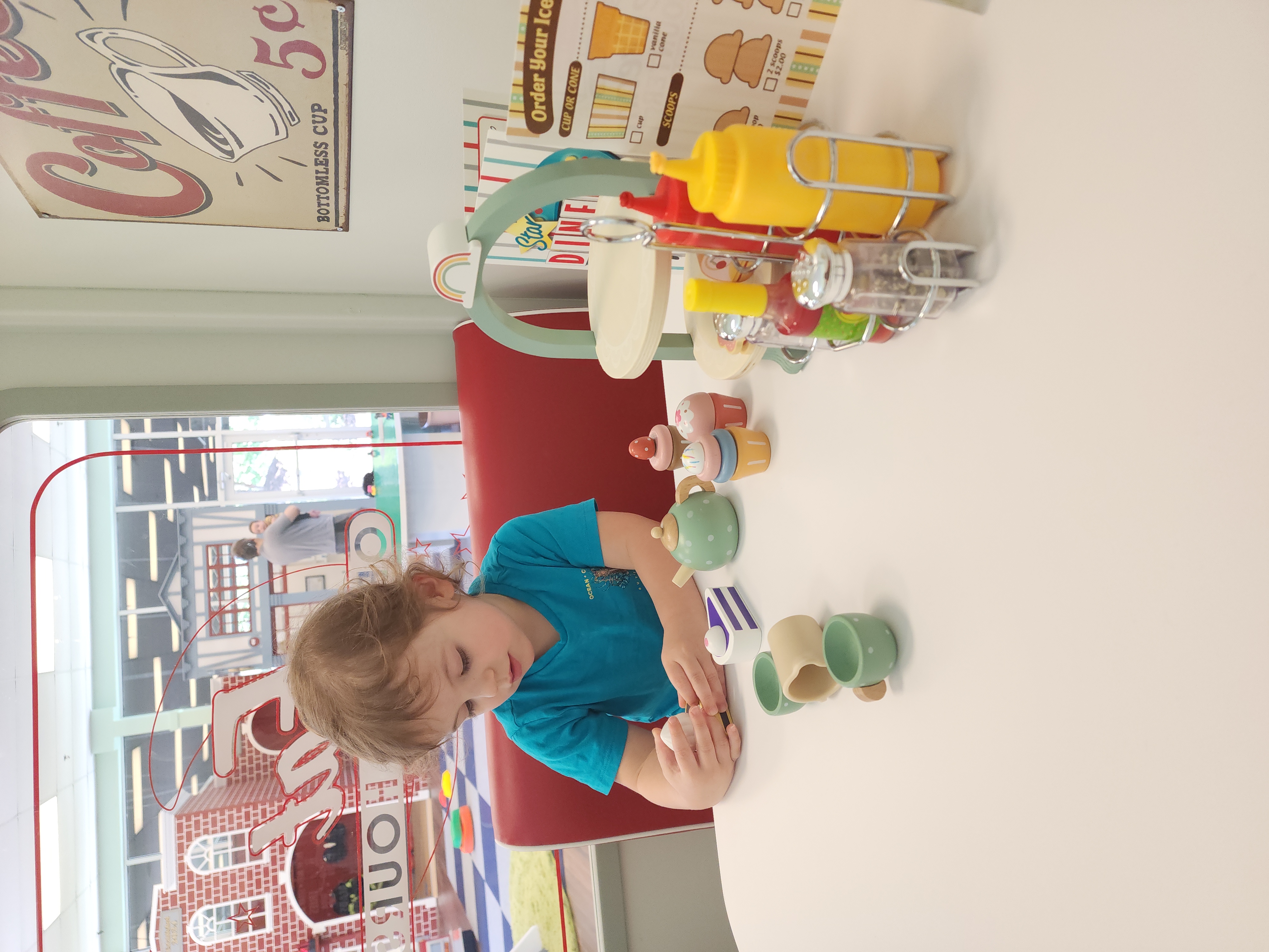 A young child plays at a pretend diner setup within Town of Fun indoor playground, arranging toy tea cups, a teapot, cupcakes, and condiments on a small white table. The playful decor includes a retro 'Coffee, 5¢' sign on the wall, and a colorful ice cream order board, with the larger playground scene visible through the diner window in the background.