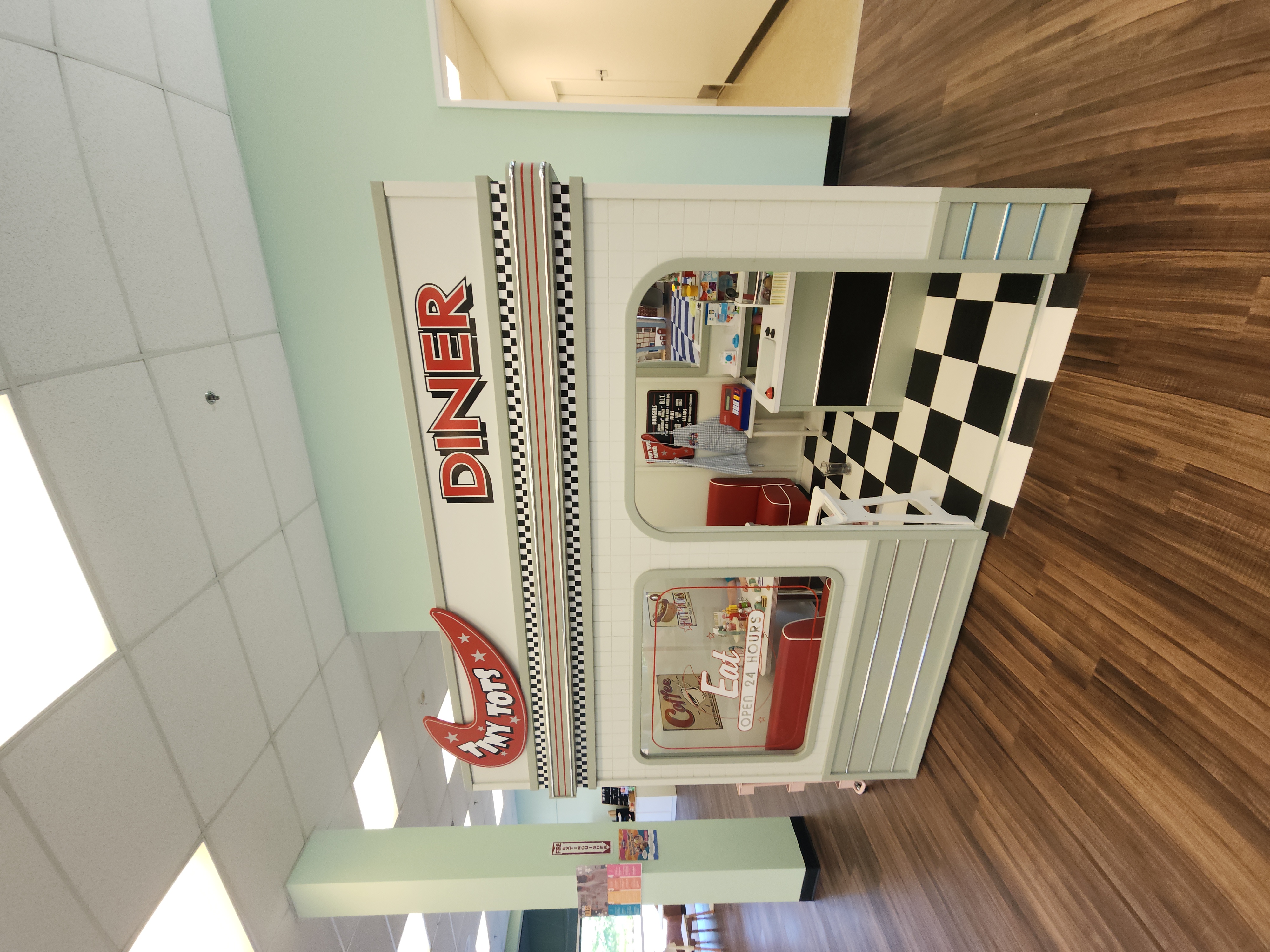 Front view of the 'Tiny Tots Diner' at Town of Fun indoor playground, featuring a vintage-inspired design with a checkerboard pattern floor, retro signage, and a small play kitchen inside. The window displays playful posters, including a 'Coffee Open 24 Hours' sign, adding to the nostalgic diner atmosphere. The light-colored walls and wooden floors of the playground surround the diner setup