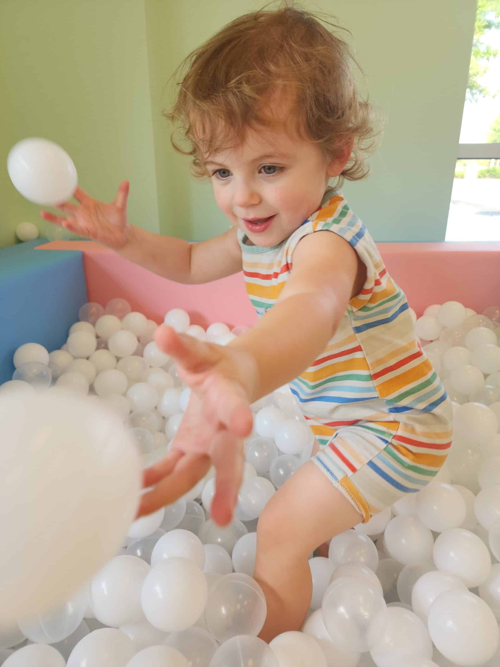 A young child with curly hair joyfully plays in a ball pit filled with white plastic balls at Town of Fun indoor playground. Wearing a colorful striped romper, the child throws a ball with an excited expression, surrounded by soft, pastel-colored walls.