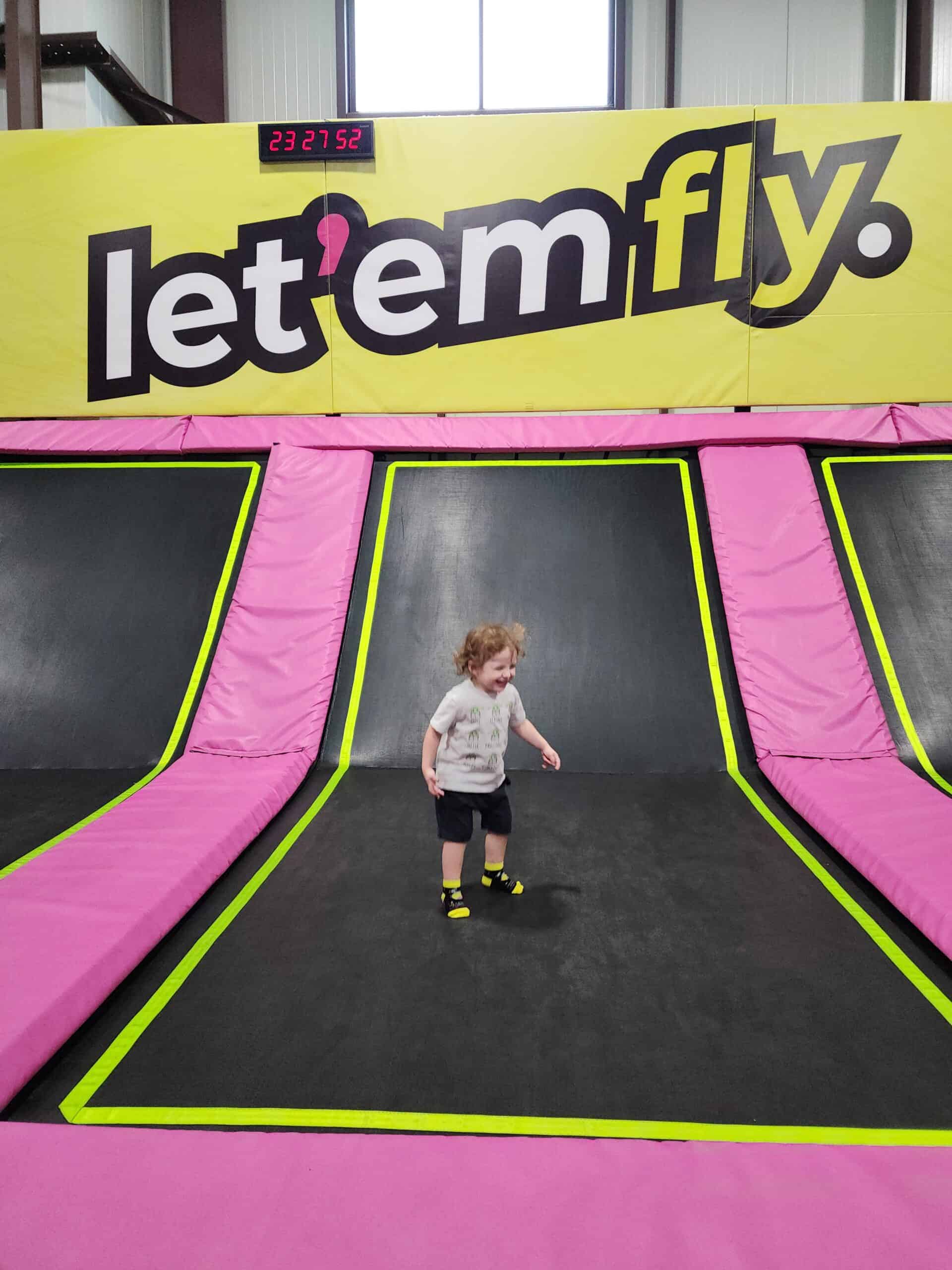 This image features a child happily bouncing on a trampoline at a facility with bold signage that reads "let 'em fly" on a yellow background. The trampoline area is framed with bright pink padding and neon green accents, ensuring both safety and vibrancy. A digital clock above the sign adds a sporty touch, reinforcing the active, energetic environment.