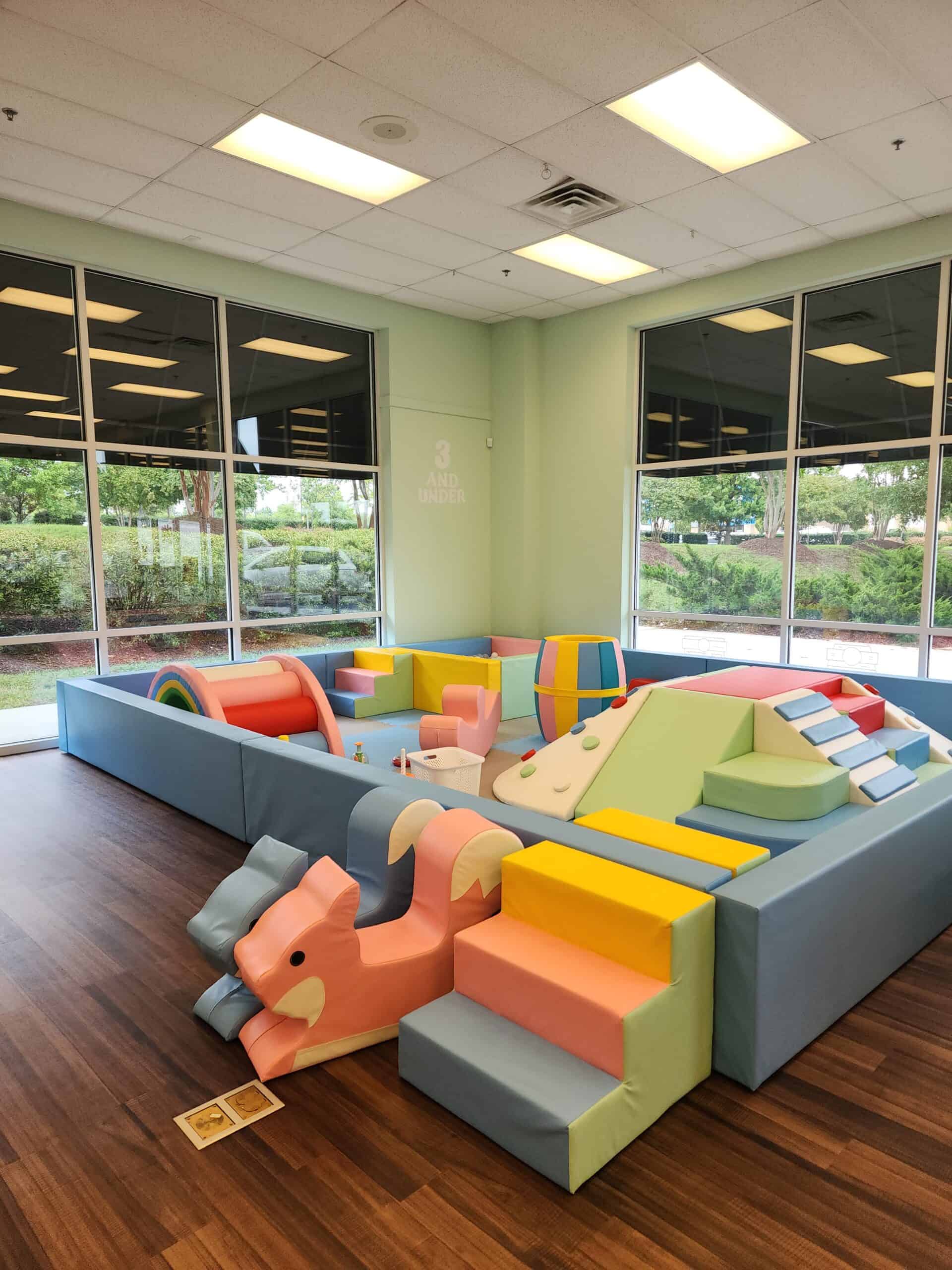 A soft play area designed for children aged 3 and under at Town of Fun indoor playground, featuring pastel-colored foam structures, including steps, slides, and climbing elements. A pink squirrel-shaped slide sits at the entrance, and a variety of ramps and soft blocks fill the enclosed space. Large windows bring in natural light, with greenery visible just outside.