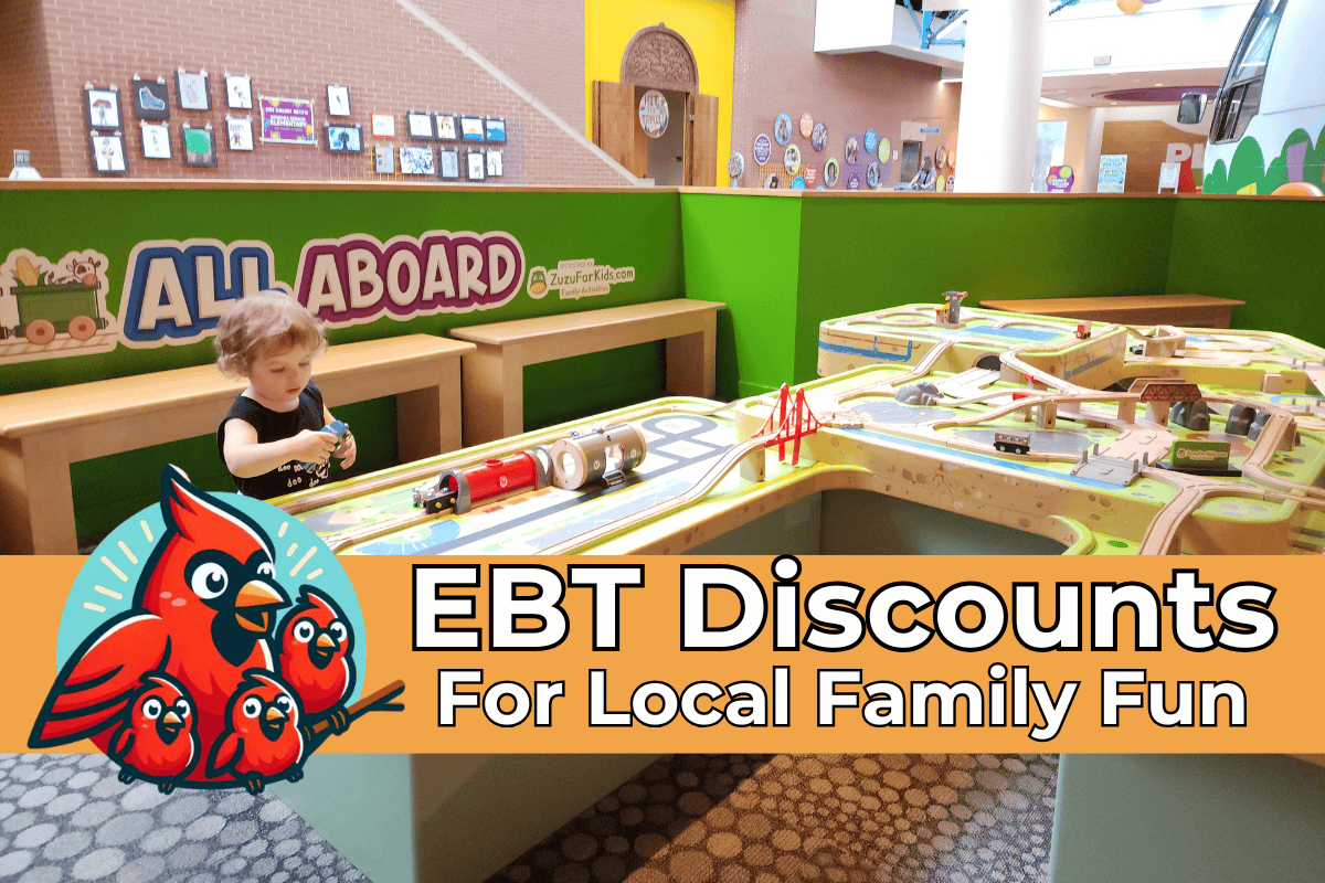 A young child plays at a colorful wooden train table inside a children’s museum or play center, with a green wall behind them that says “ALL ABOARD” in bold letters. A graphic overlay in the foreground features red cartoon cardinals and reads, “EBT Discounts For Local Family Fun.”